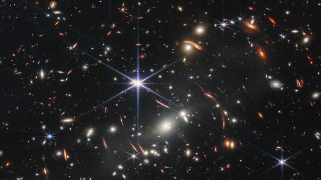 Tusenvis av små galakser. Fargene deres varierer. Noen er nyanser av oransje, mens andre er hvite. De fleste vises som uklare ovaler, men noen få har distinkte spiralarmer. Foran galaksene er det flere stjerner. De fleste ser blå ut, og de lyse stjernene har diffraksjonsspor som danner en åttepunktet stjerneform. Det er også mange tynne, lange, oransje buer som bøyer seg rundt sentrum av bildet. Foto.