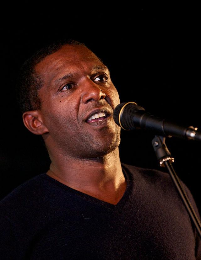The British poet Lemn Sissay is talking into a microphone.  He's a coloured man, dressed in a black sweater. Black background. Photo.