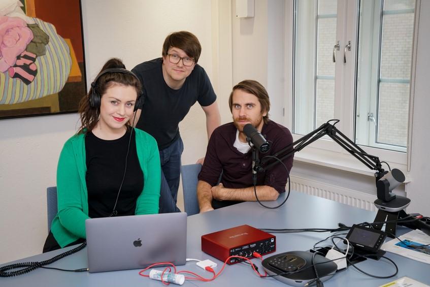 Tre personer sitter ved en datamaskin foran en studiomikrofon.  Foto.