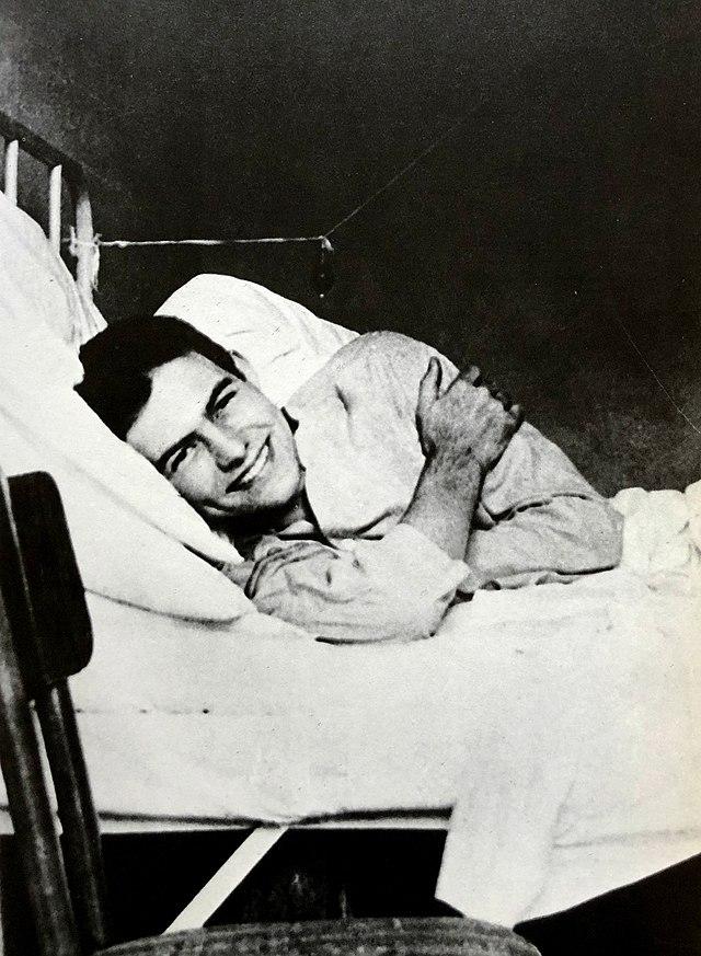 Photo: We see a young man. He is lying on his side in a hospital bed. He is smiling towards the camera. 