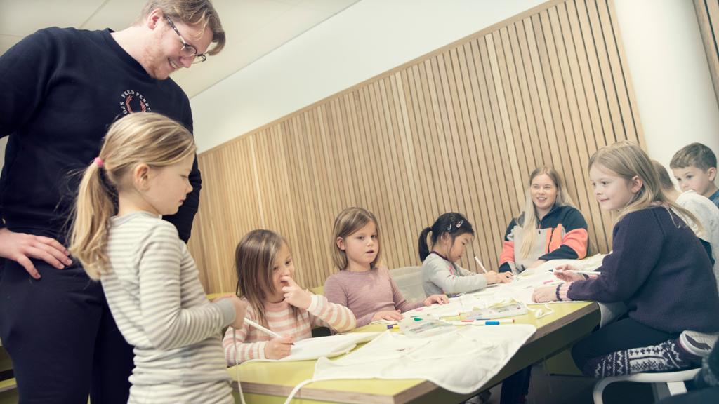 To voksne har samlet ei gruppe barn rundt et bord.  Barna maler og tegner. Foto.