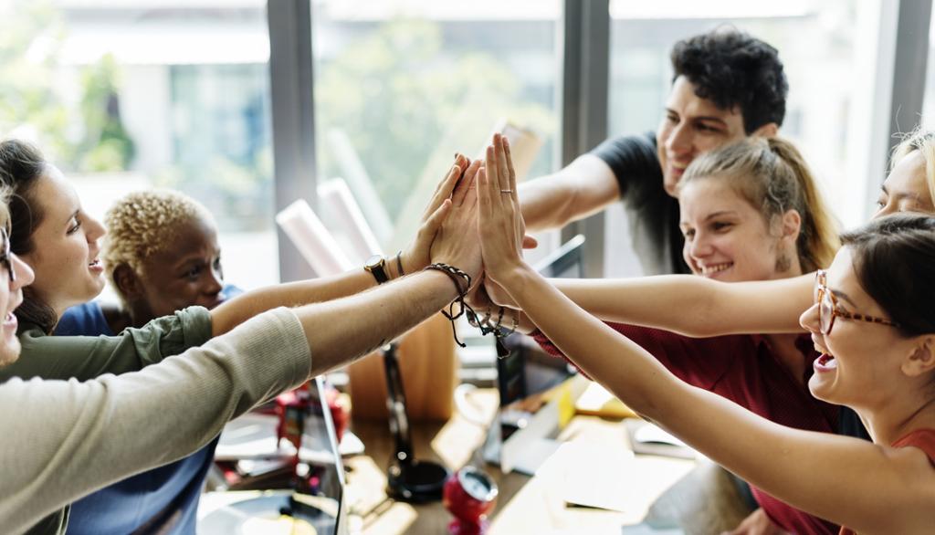 En gjeng unge voksne smiler og gir hverandre en "high five" over arbeidsbordet. Foto. 