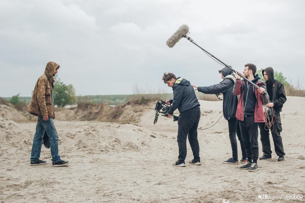 En mann med dongeribukse og en brun jakke går på sand. Foran han står et kamerateam som filmer han og tar opp lyd med en lydbom. Foto.