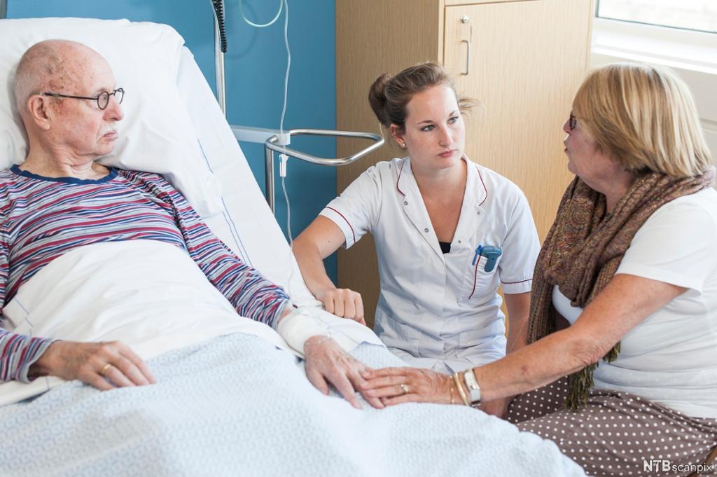 Eldre mannlag pasient og kvinneleg pårørande i samtale med kvinneleg sjukepleiar. Mannen ligg i ei sjukeseng, kvinna held handa hans. Foto.