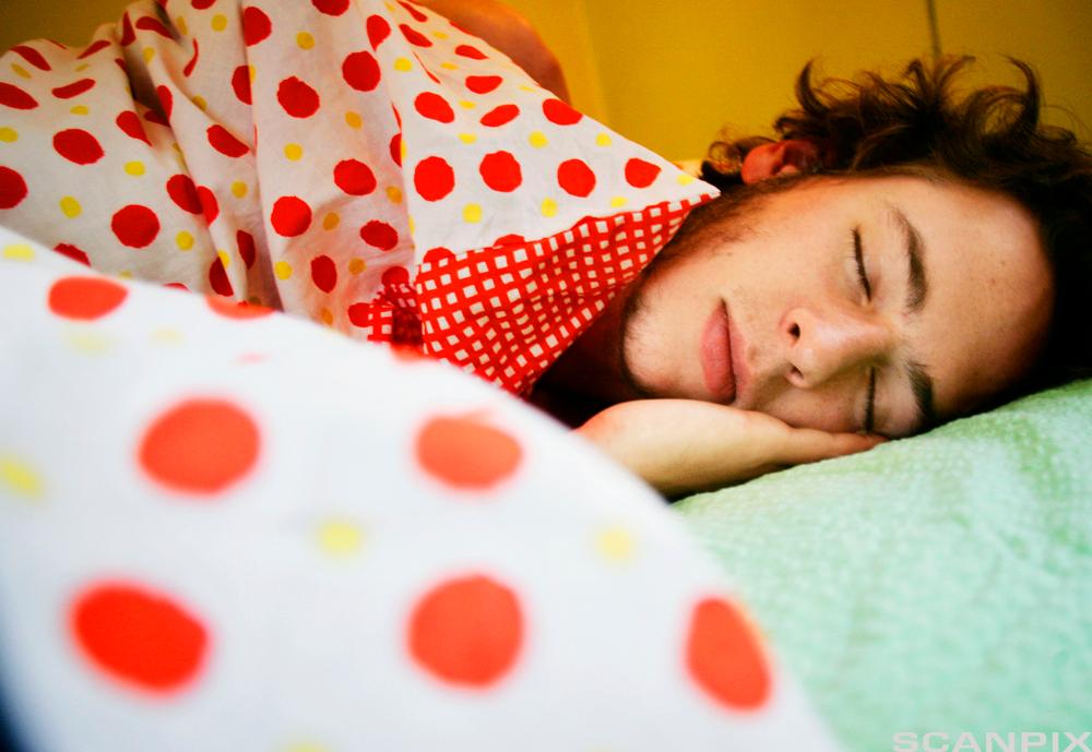 A sleeping teenage boy. He has a green pillow, and a duvet with orange and yellow dots. Photo.
