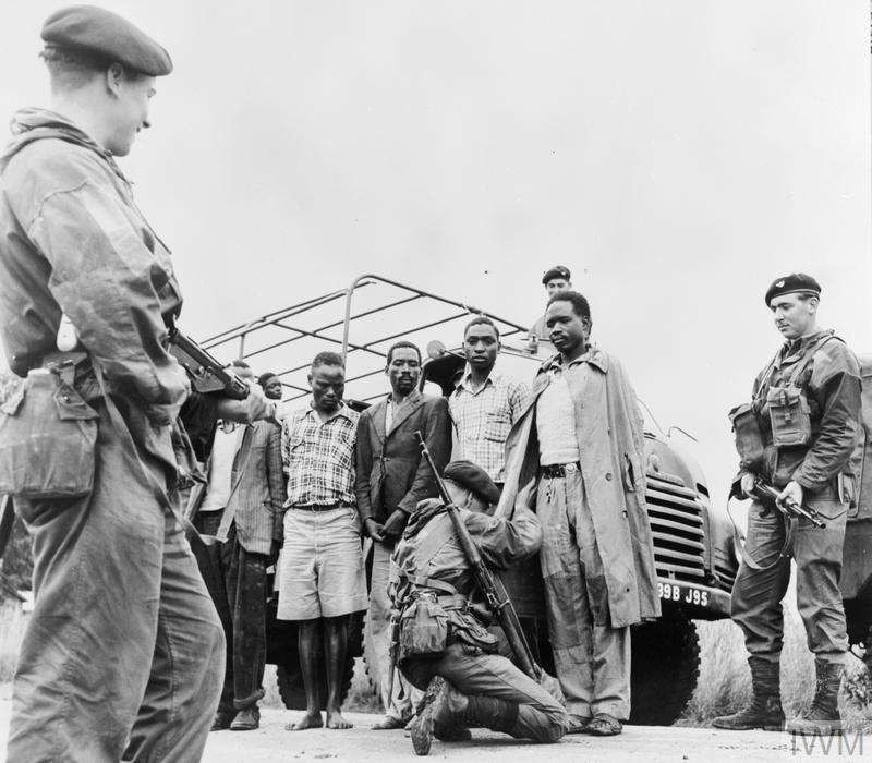 Photo: We see one soldier pointing a weapon at a group of Black men. Another soldier is holding his gun with both hands, pointed away from the men. One soldier is on his knees searching one of the Black men. The soldier has a gun on his back. In the background, there is a lorry. 