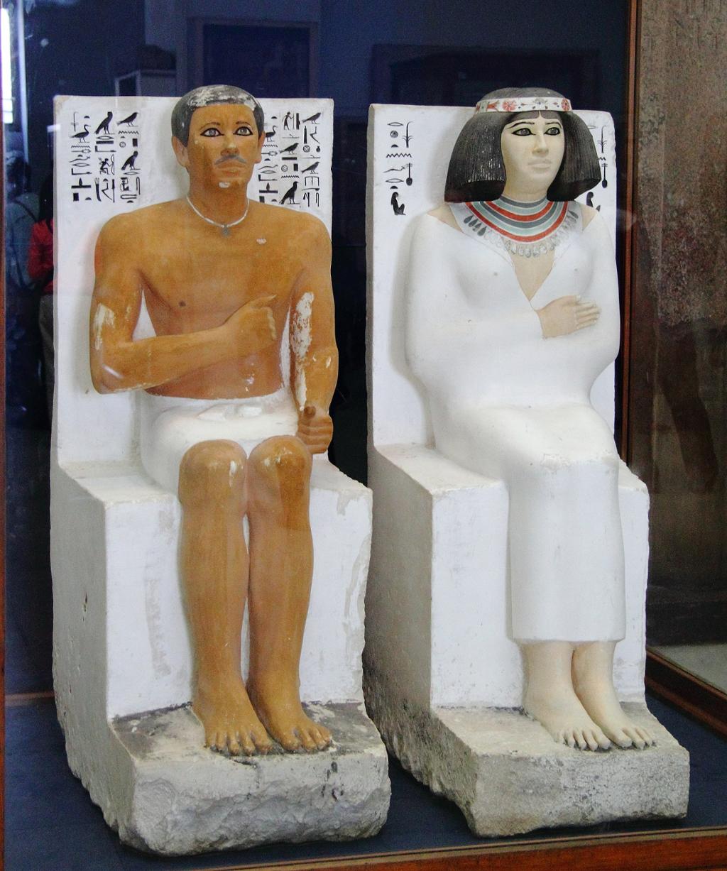 Skulpturer av et egyptisk par som sitter med høyre hånd opp mot brystet og beina rolig ved siden av hverandre. De ser rett fram. Foto.