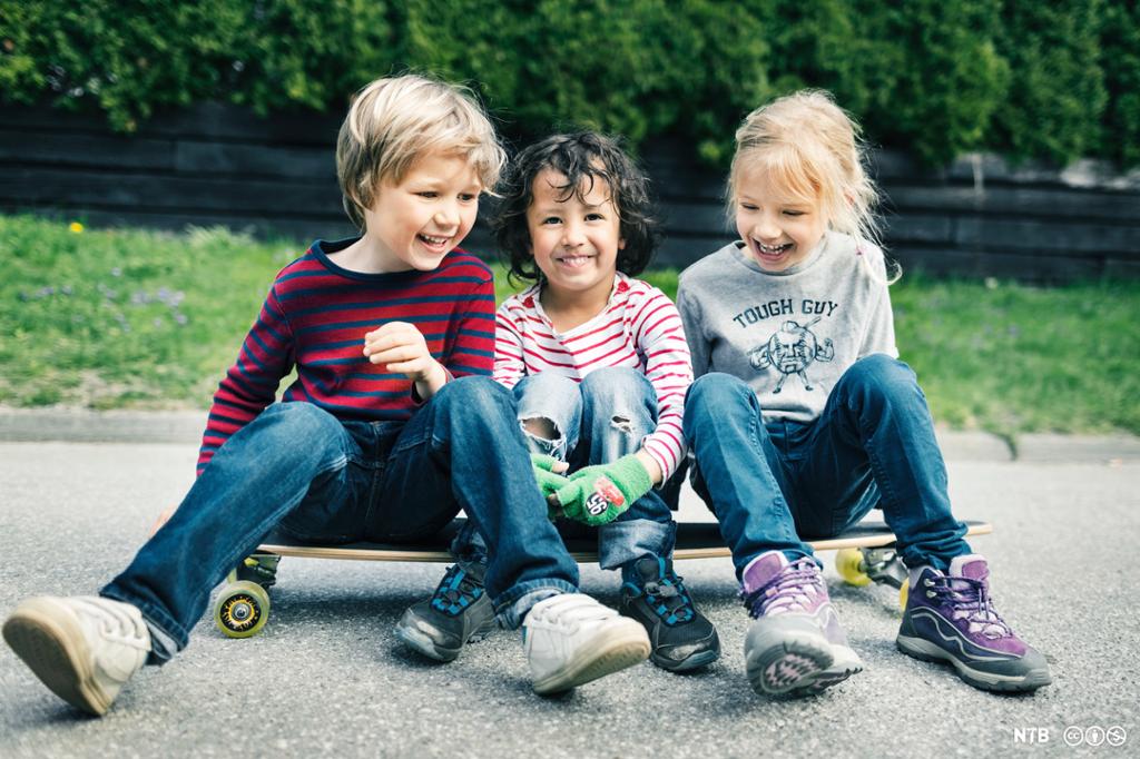 Tre smilande barn sit på eit skateboard. Foto. 