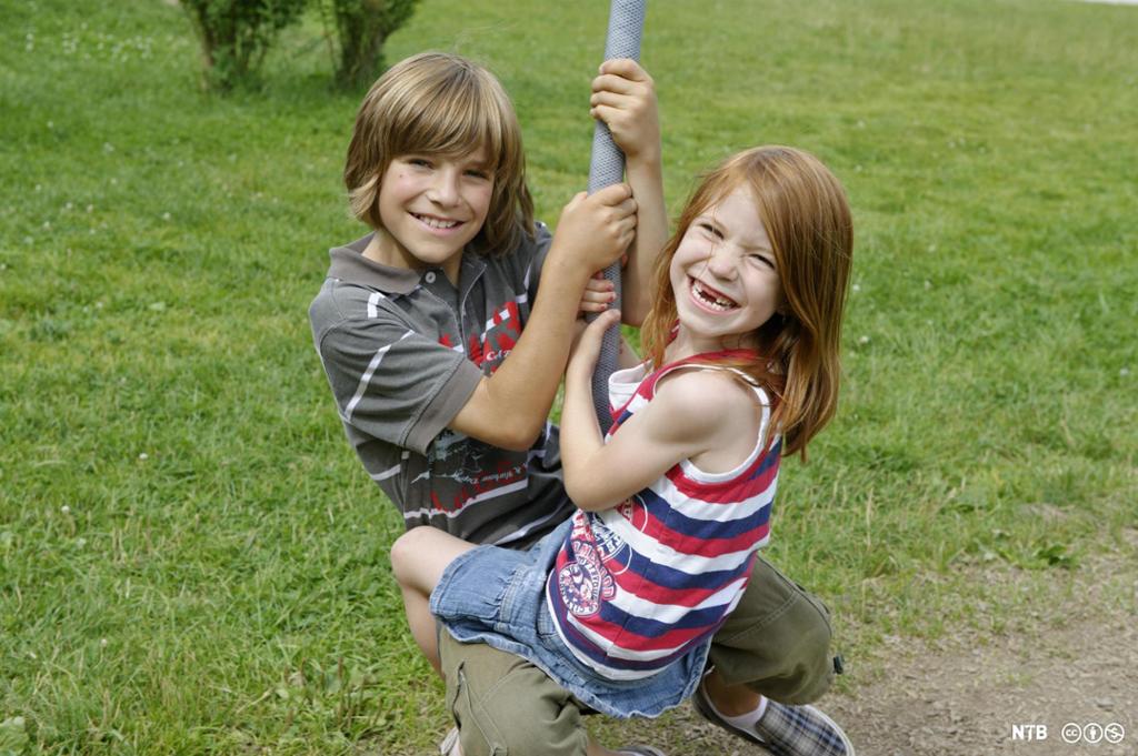 En gutt og ei jente sitter mot hverandre og holder seg fast i et tau mens de ser mot kamera og smiler. Foto.  