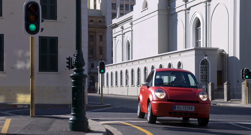 En liten, rød elbil fra det tidligere norske bilmerket Think kjører i ei gate. På bildet er et veikryss med grønt trafikklys og oppmalte felt i veien omgitt av hvite bygninger. Foto. 