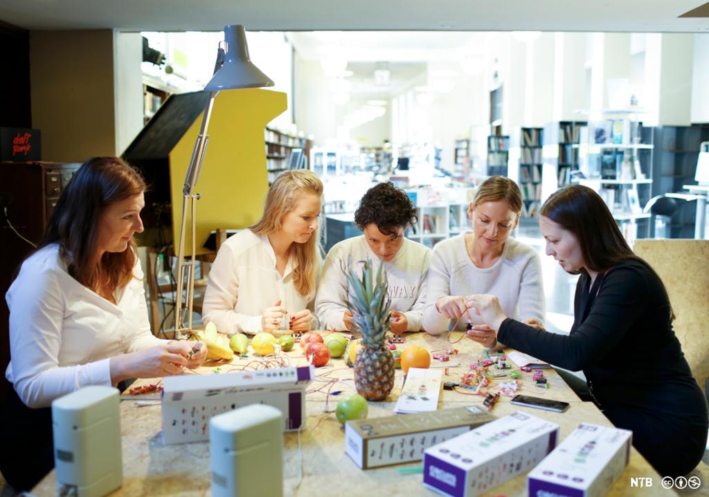Fem kvinner sitter rundt et bord og eksperimenterer med teknologi og koding. Bordet er fullt av ledninger, frukt og esker. Foto. 