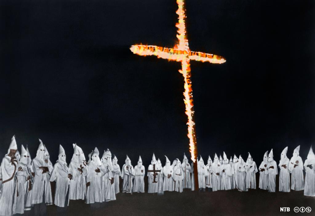 En ring av personer i hvite, heldekkende drakter samlet rundt et brennende kors. Foto.