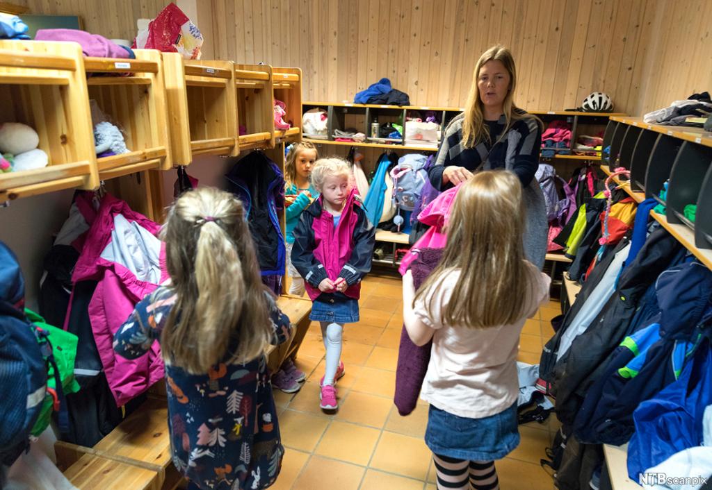 En voksen og fire jenter i garderoben på en skole. Den voksne hjelper barna med å finne fram yttertøy. Foto.