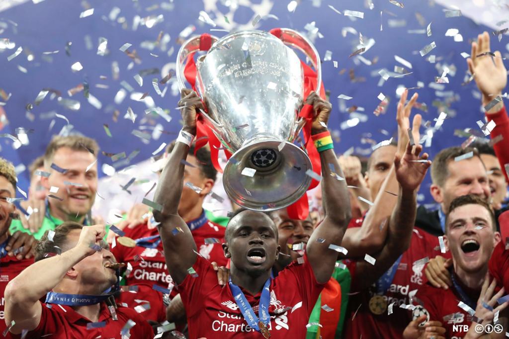 En fotballspiller løfter en trofe over hodet med jublende mennesker rundt. Spillerne er kledd i røde fotballskjorter. Konfetti svever i luften overalt. Foto.