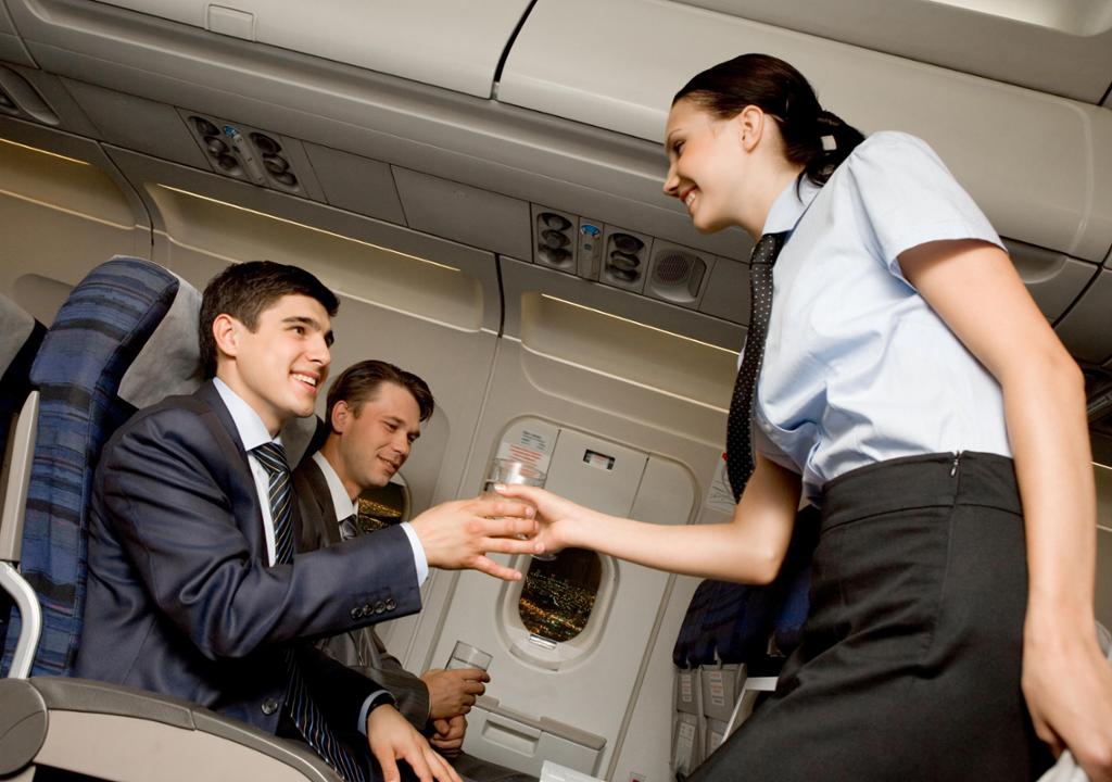 Flyvertinne serverer et glass vann til flypassasjer. Passasjeren er kledd i dress og slips og ser smilende opp på flyvertinnen, som smiler tilbake. Foto.