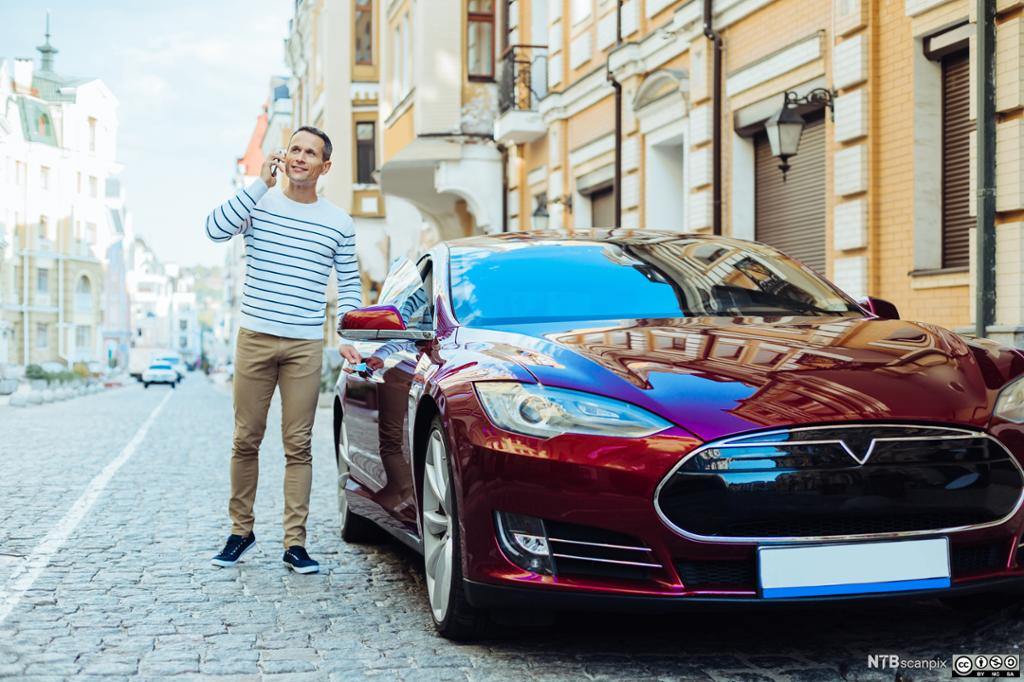 En mann snakker i mobilen på vei inn i den røde Tesla-sportsbilen sin. Foto.