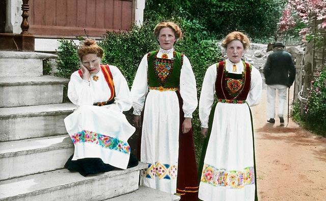 Tre kvinner i bunad ved ei trapp. Den ene sitter på trappa med hodet lent i hånda, mens de to andre står ved siden av. I bakgrunnen går en mann med stokk. Foto.