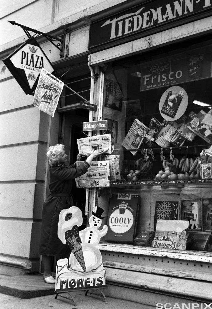 Bilde av en kvinne som plasserer Aftenposten i et avisstativ utenfor en kiosk 19. juni 1962.