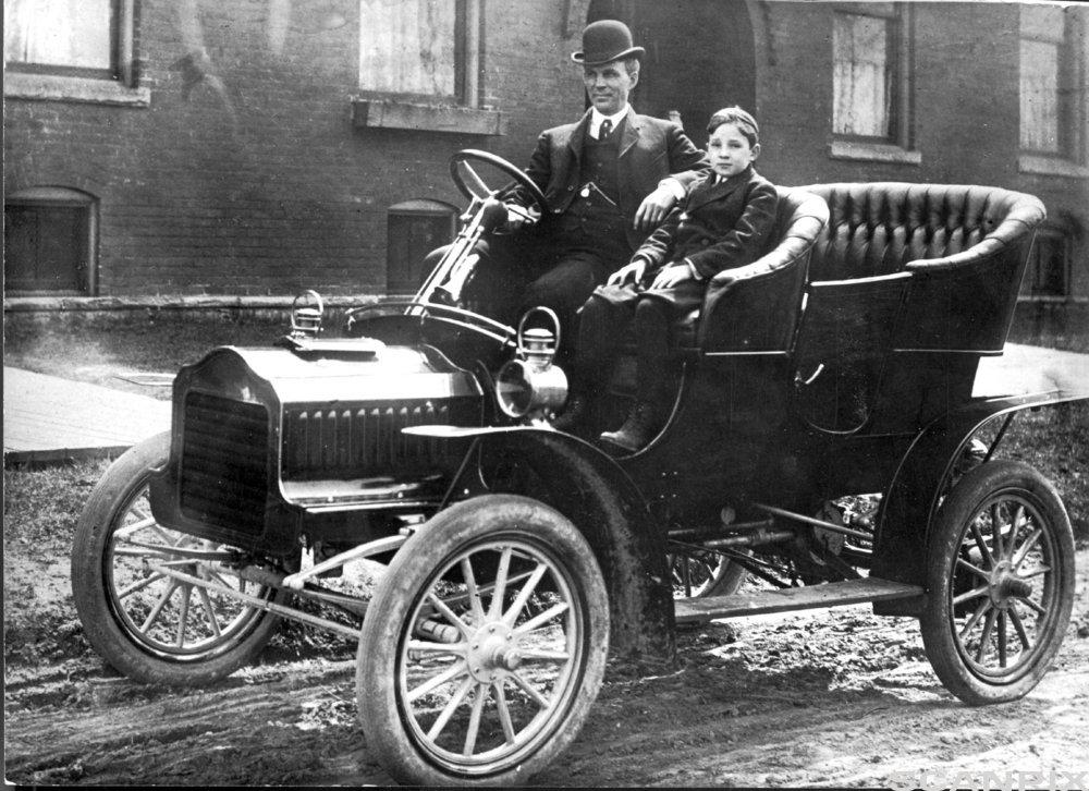 En mann og en gutt sitter i en bil fra begynnelsen av 1900-tallet. Svart-hvitt-foto. 