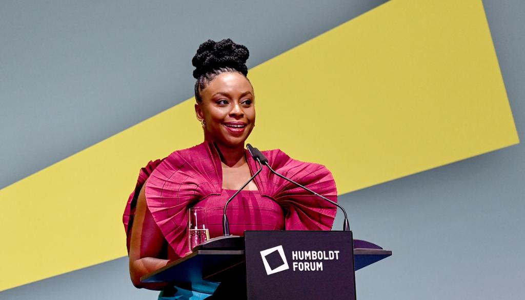 Den unge forfattaren Chimamanda Ngozi Adichie står på talarstolen. Ho har på seg ein sjokkrosa bluse. Foto.
