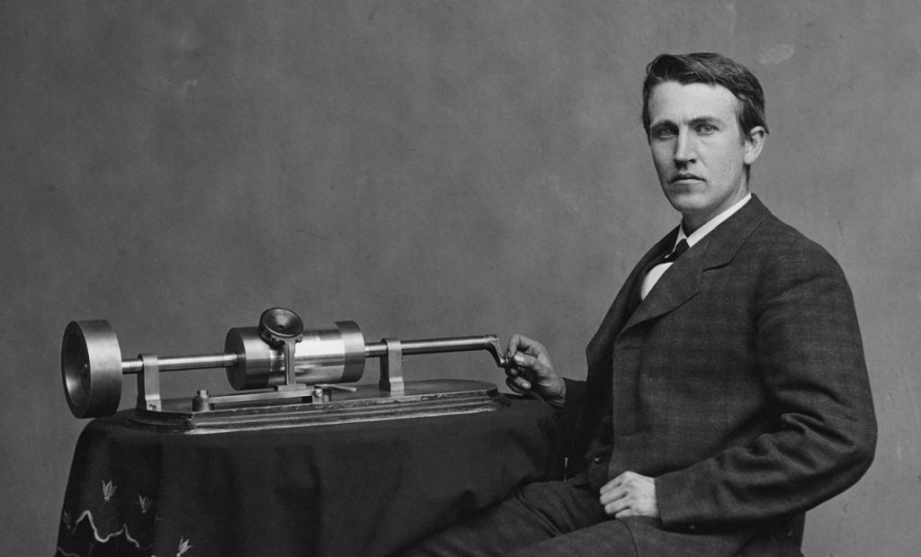 Photo: Thomas Alva Edison sitting next to a phonograph placed on a table with an embroidered table cloth. He is wearing a suit and leather shoes. 