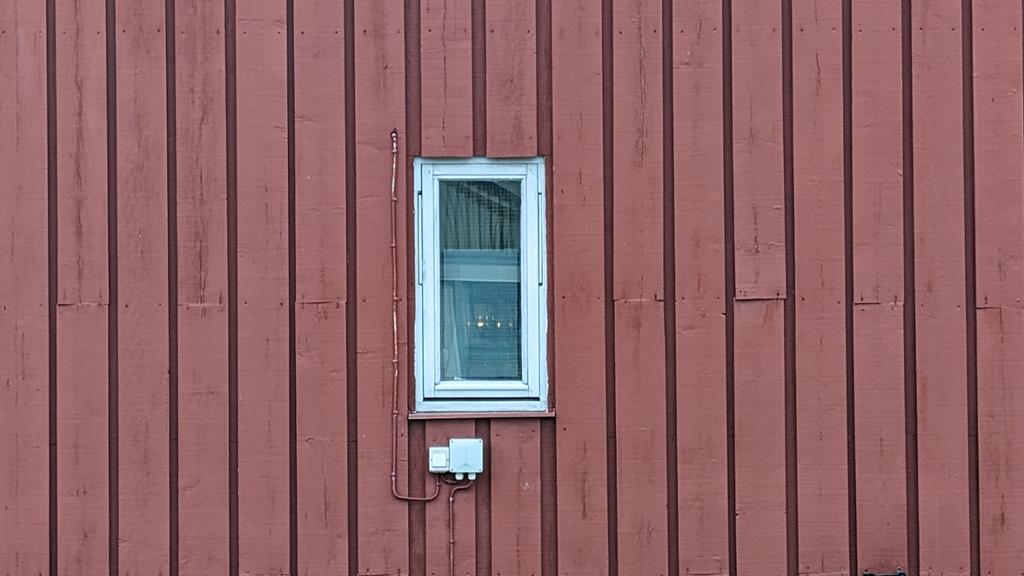 En rød husvegg med et smalt vindu. Overliggerne på siden av vinduet ligger relativt tett. Over og under vinduet ligger ikke overliggerne like tett. Foto.