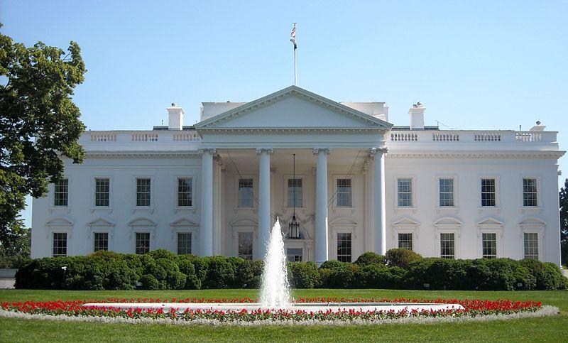 Photo: The White House in Washington D.C. We see a white buildings with columns. There is a flag on the roof. There is a fountain in front of the house. 