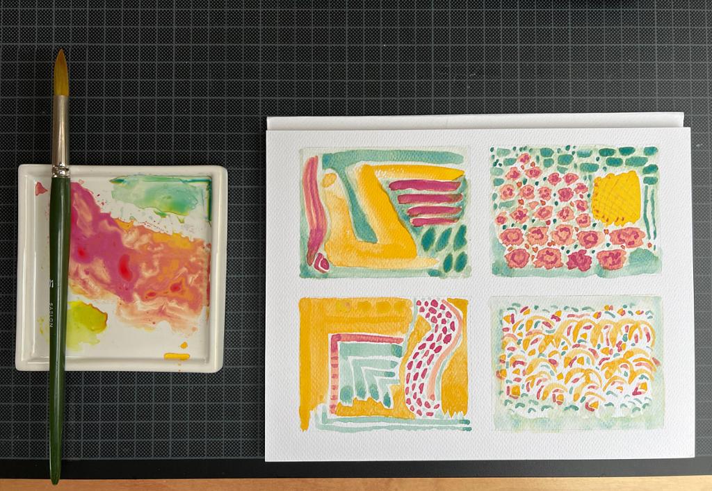 En malerkost, en blandepalett og ei blokk med akvarellpapir hvor det er malt abstrakte former i rødfiolett, guloransje og blågrønt. Foto.