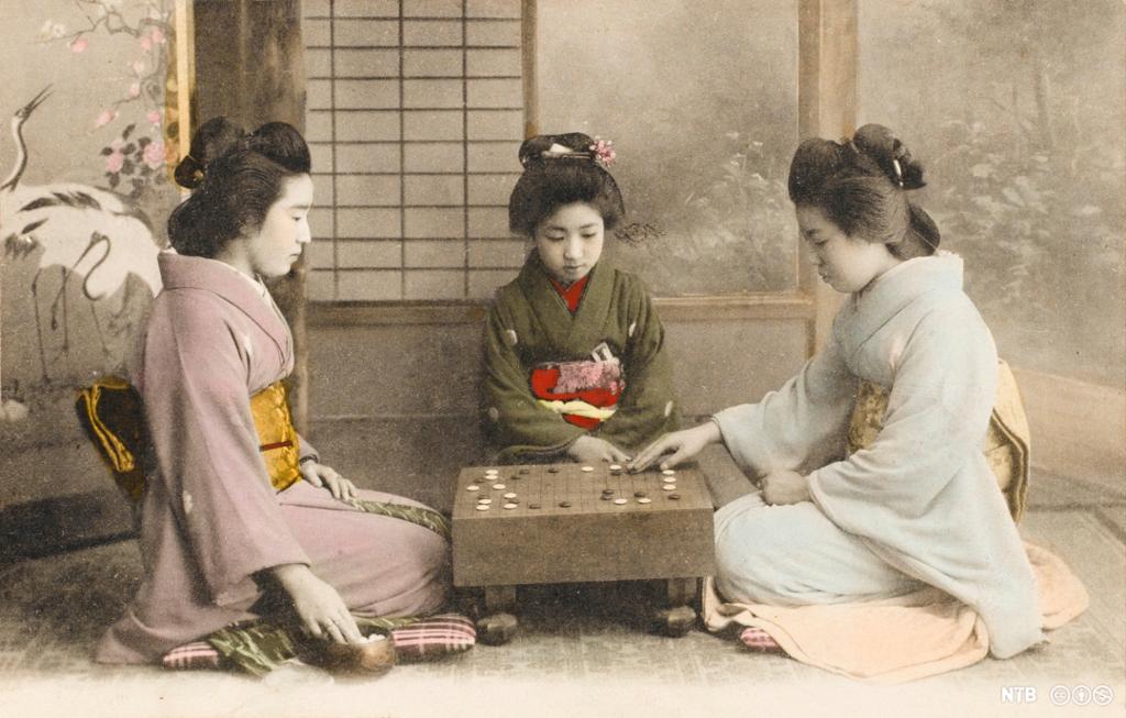 Tre japanske jenter i kimono sit rundt eit lite firkanta bord med svarte og kvite spelebrikker på. Foto.
