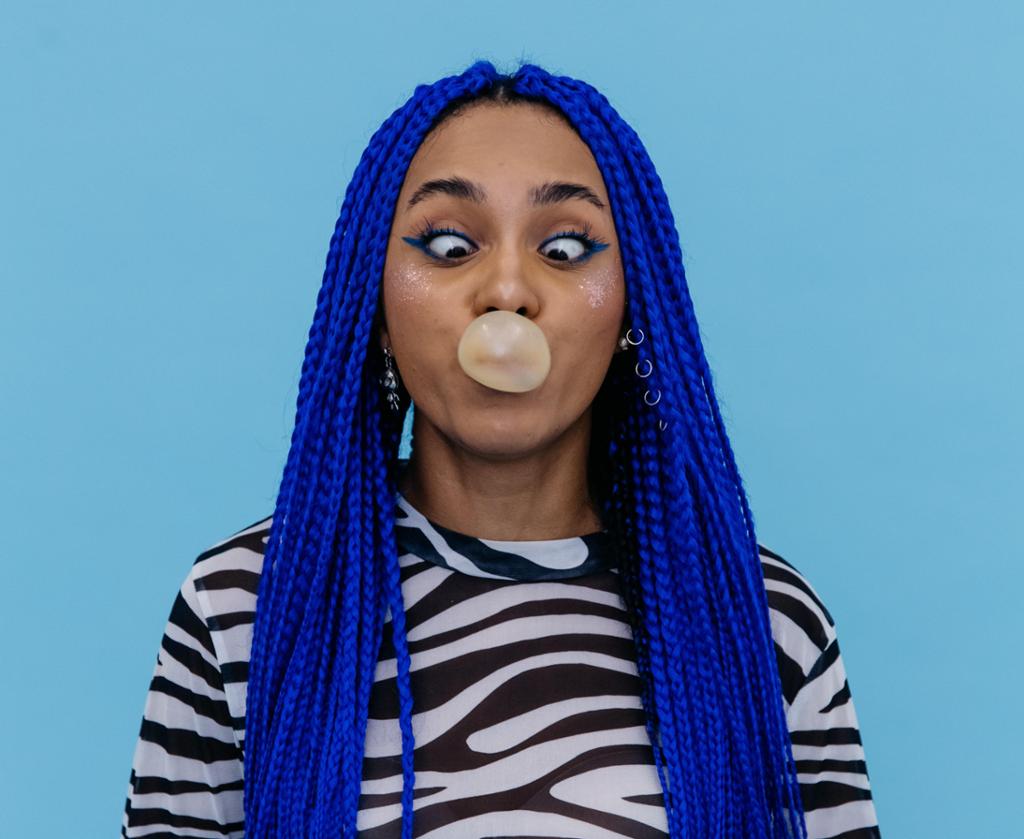 Ei ung kvinne med lang, blått hår blåser ei tyggegummi-boble og ser ned på boblen hun blåser. Foto.