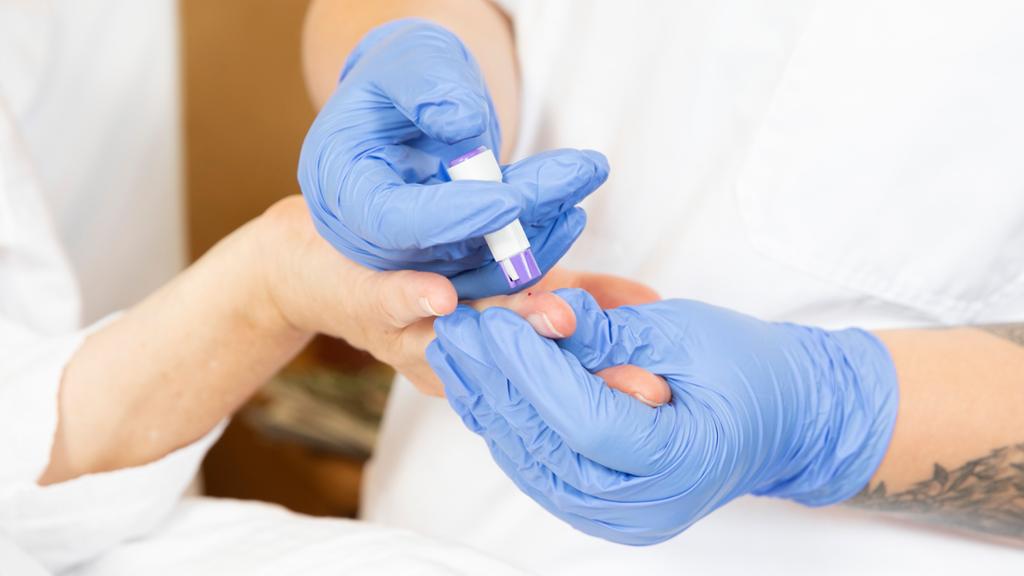 En person med hansker tar blodprøve fra fingeren til en pasient. Foto.