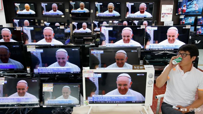 Mann sitter i butikk med mange tv-er. Paven er på tv-skjermene. Foto.