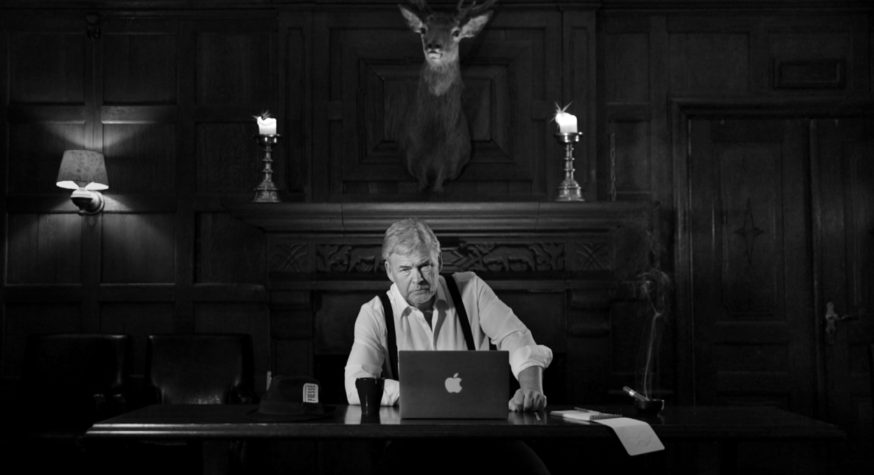 Eldre herre i hvit skjorte sitter bak et gammeldags skrivebord med en alvorlig mine. Foto.