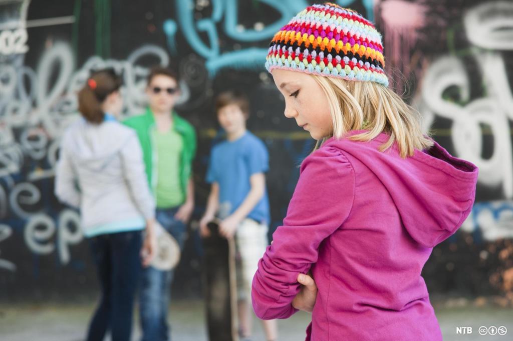 Tre tenåringer står sammen foran en graffitivegg. Ei ung jente står alene. Foto.