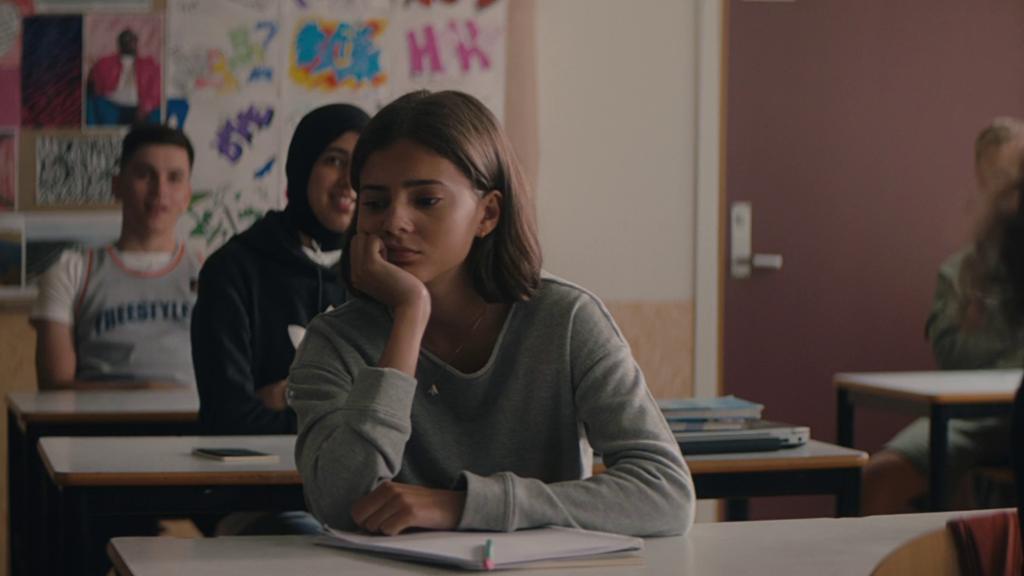 En jente sitter med hodet i hånden og tenker ved en pult i et klasserom. Foto.