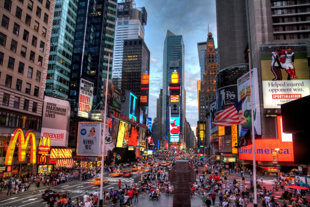 Times Square i New York, med mye trafikk og et yrende folkeliv. Gatene er omkranset av høyhus og skyskrapere, og på fasadene henger store, opplyste reklameskilt. Foto.