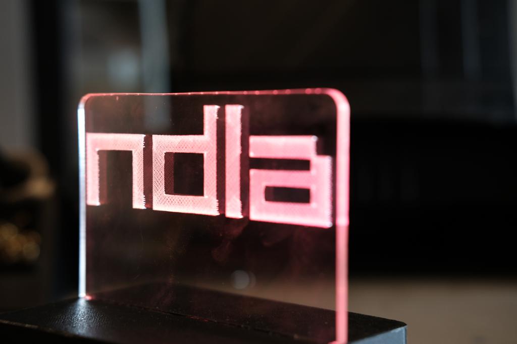 Et bordskilt i gjennomsiktig pleksiglass, med opplyst NDLA-logo. Foto.