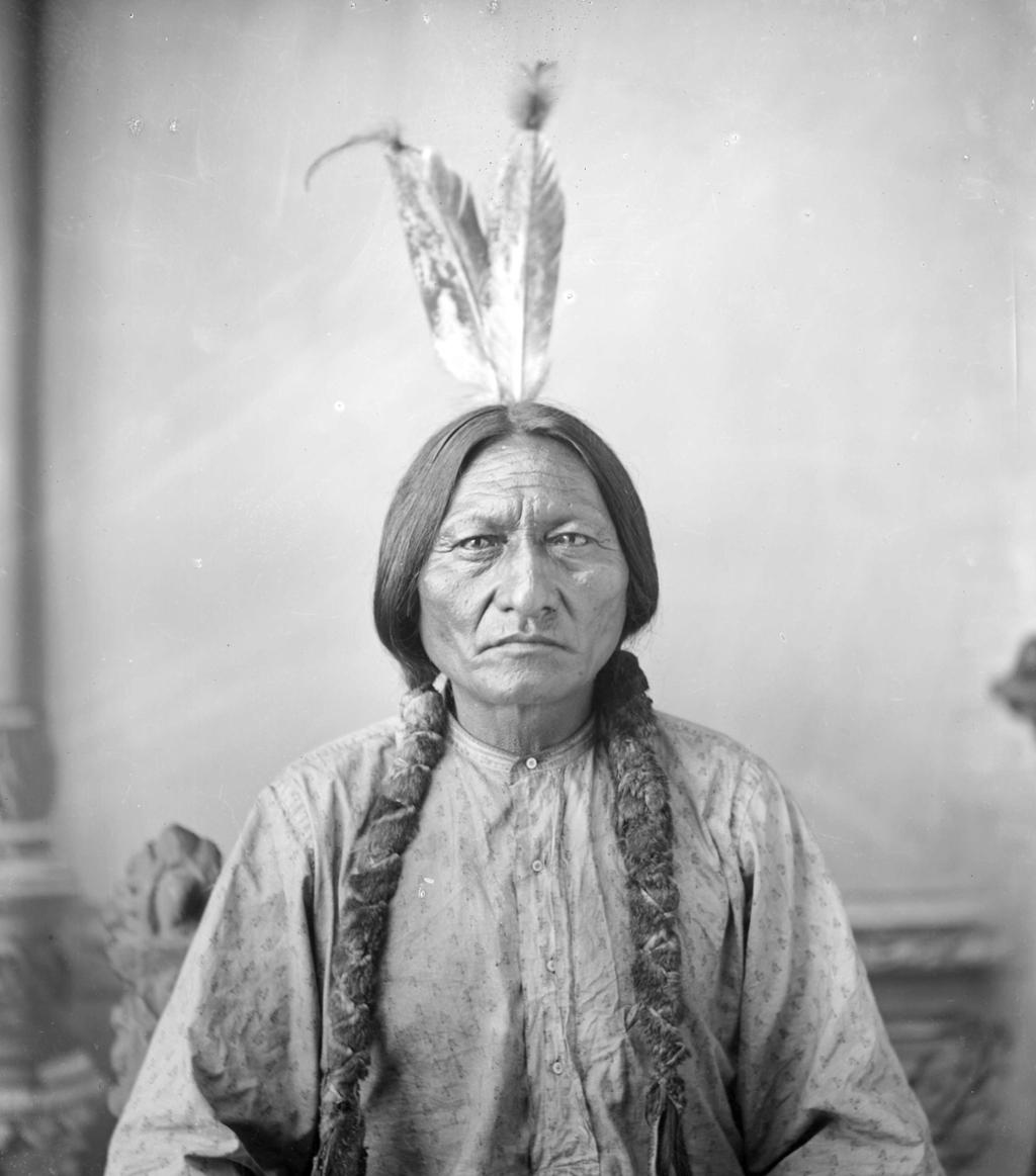 Photo: Portrait of Sitting Bull. We see an older, Native American man. His hair is in two thick braids. He wears a patterned shirt and has two feathers in his hair. 