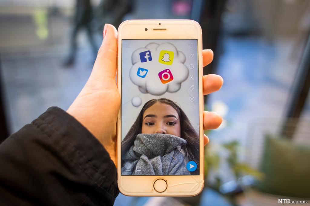 Ei hånd holder en mobiltelefon med bilde av ei jente som har logoer fra ulike sosiale medier i ei tenkeboble over hodet. 