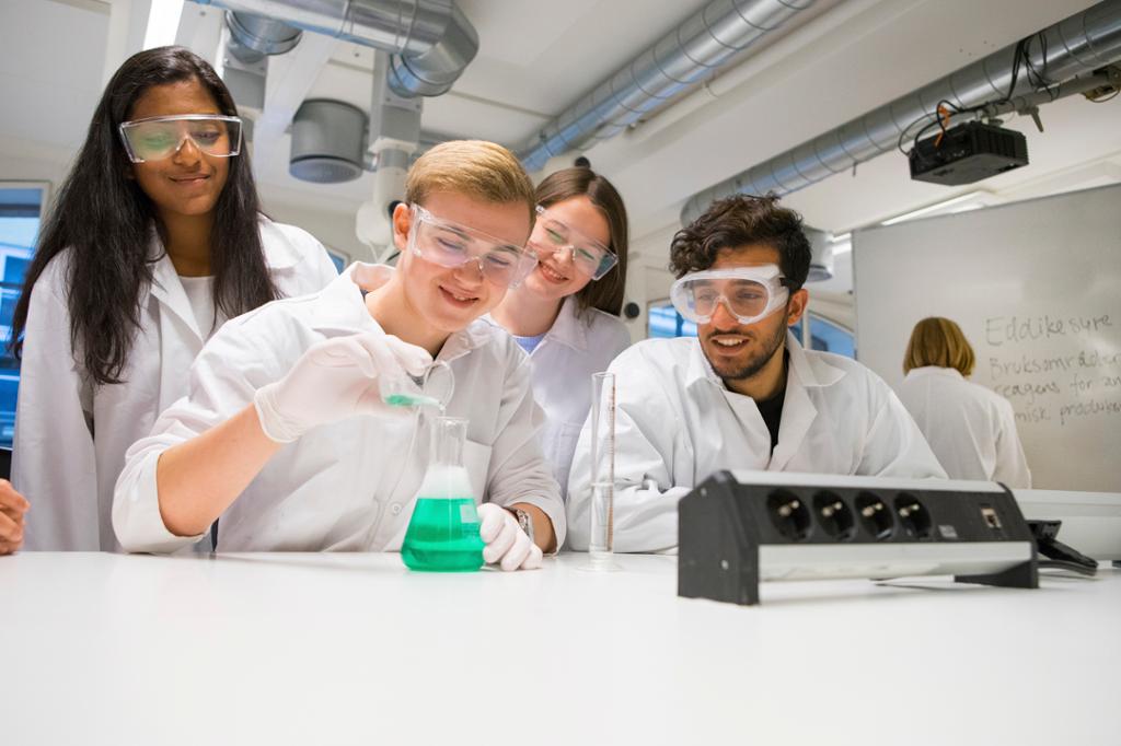 Fire elever med lærer i bakgrunnen gjennomfører kjemisk eksperiment med grønn væske på naturfagslab. De har på seg laboratoriefrakker og beskyttelsesbriller, og eleven som blander væsker, har på seg engangshansker. Foto.