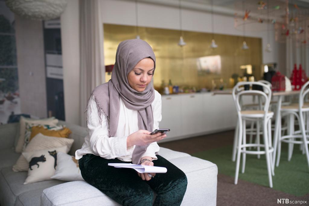 Ung kvinne med hijab tekstar på ein mobiltelefon. Ho sit på ein sofa. I bakgrunnen er eit kvitt kjøkken. Foto.