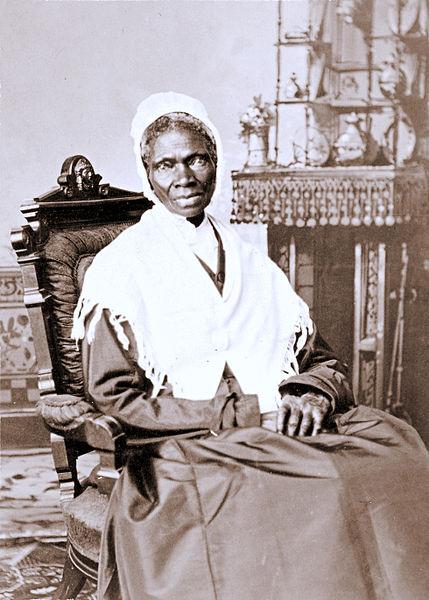 A black and white photo of Sojourner Truth, an old, Black woman sitting in a chair. She wears a dark dress and a white shawl and she has a white bonnet on her head. She's looking straight into the camera. Probably from the 1880s.    