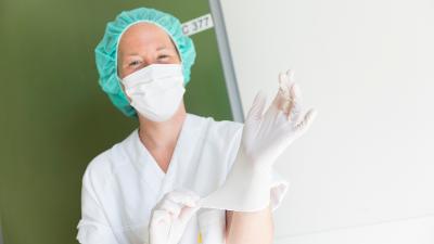 Sykepleier med munnbind og hårnett tar på seg engangs hansker. Foto.