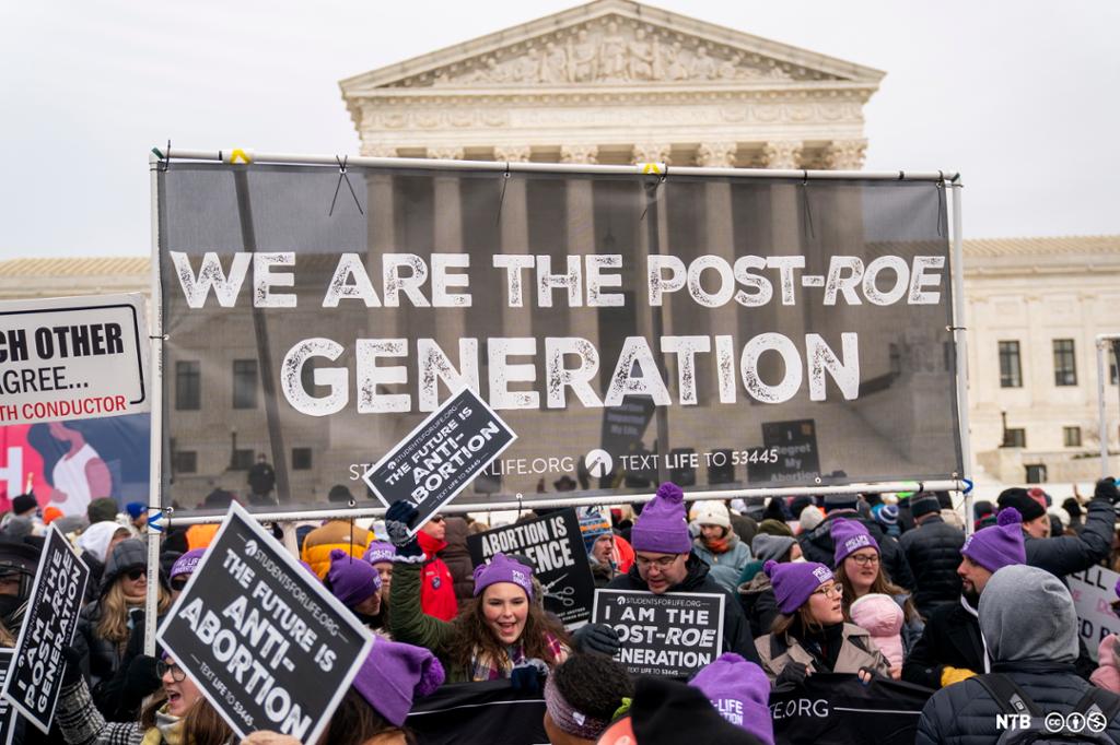 Photo: We see the US Supreme Court building in the background. In the foreground is a large banner with the words "WE ARE THE POST-ROE GENERATION". There are a number of protesters wearing purple hats, holding posters saying "THE FUTURE IS ANTI-ABORTION". 