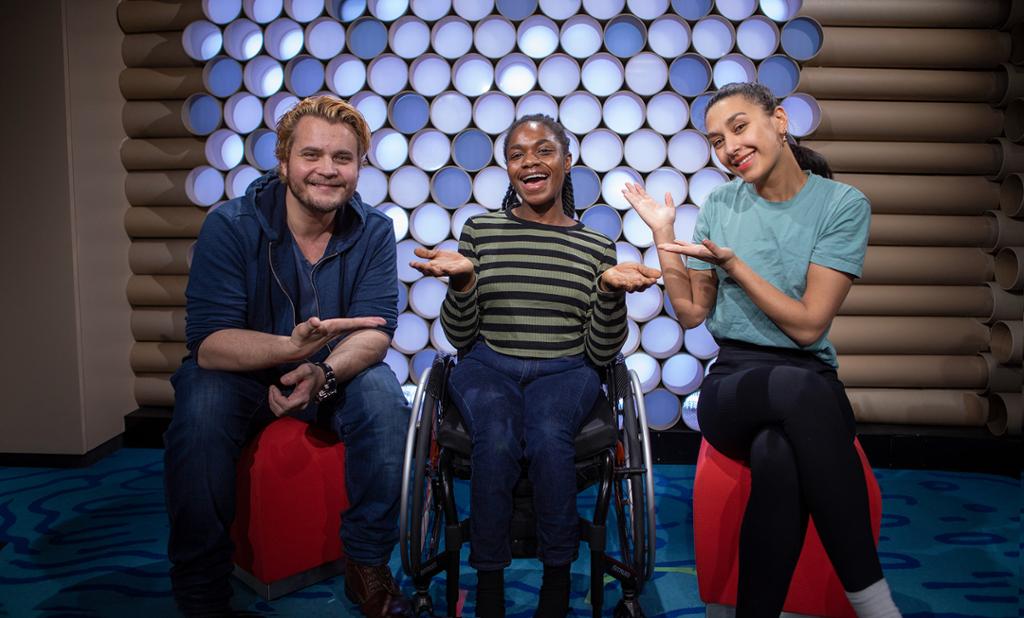 Tre unge programledere sitter i et TV-studio. Bjørn Alexander og Selda Ibrahim sitter på krakkar på hver sin side av Salamatu "Sally" Kamara, som sitter i rullestol. Foto.