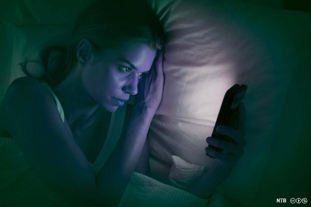 Ung kvinne ser på mobiltelefon i senga. Foto.
