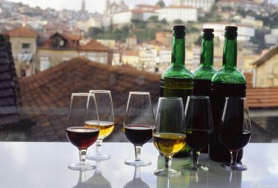 Flere glass med drikke med ulike brunfarger foran tre grønne vinflasker. Utsikt over en by i bakgrunnen. Foto.