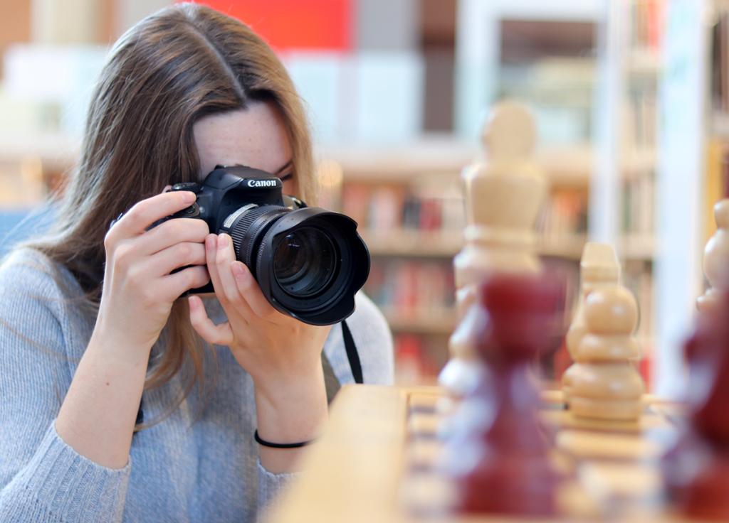 Kvinne fotograferer sjakkbrikker. Kameraet er i fokus. Sjakkbrikker i forgrunnen er ikke i fokus. Bokhyller i bakgrunnen har liten dybdeskarphet. Foto.