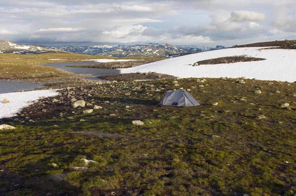 Et grått telt er satt opp på et flatt område med gress og mose. Rundt er det store flekker med snø, i bakgrunnen er det fjell så langt øyet kan se. Foto