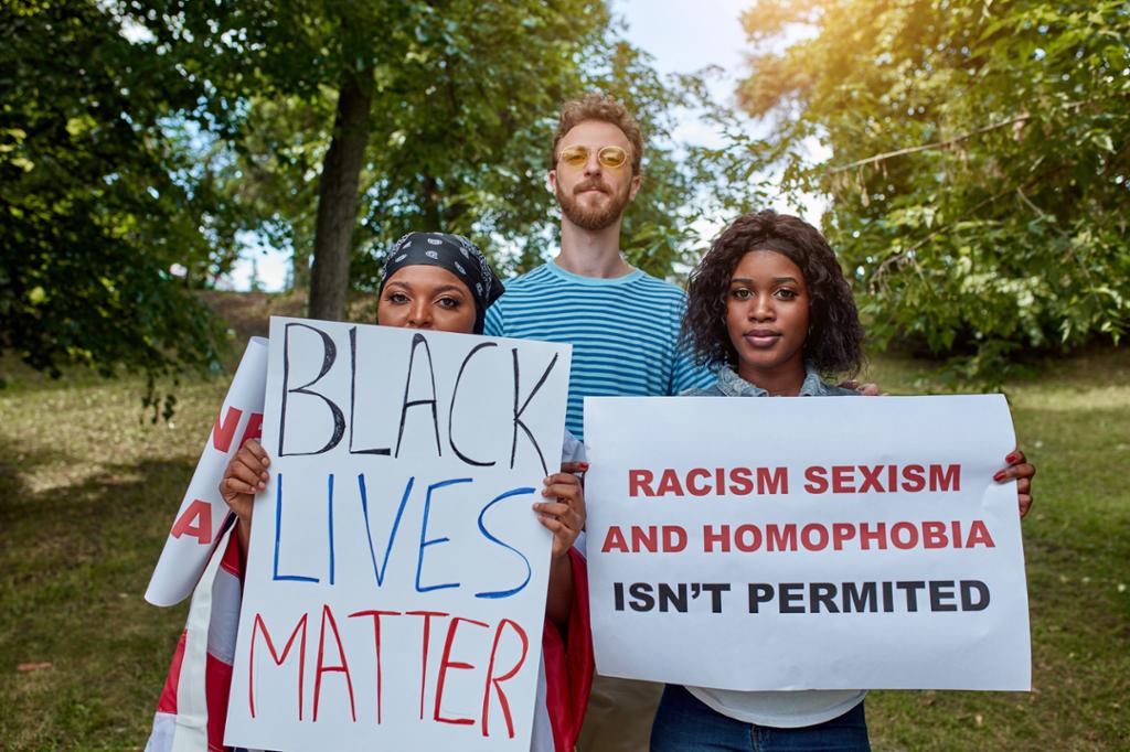 Tre mennesker står sammen, to holder plakater med slagord på engelsk: "Black lives matter" og "Racism, sexism, and homophobia isn't permitted". Foto.